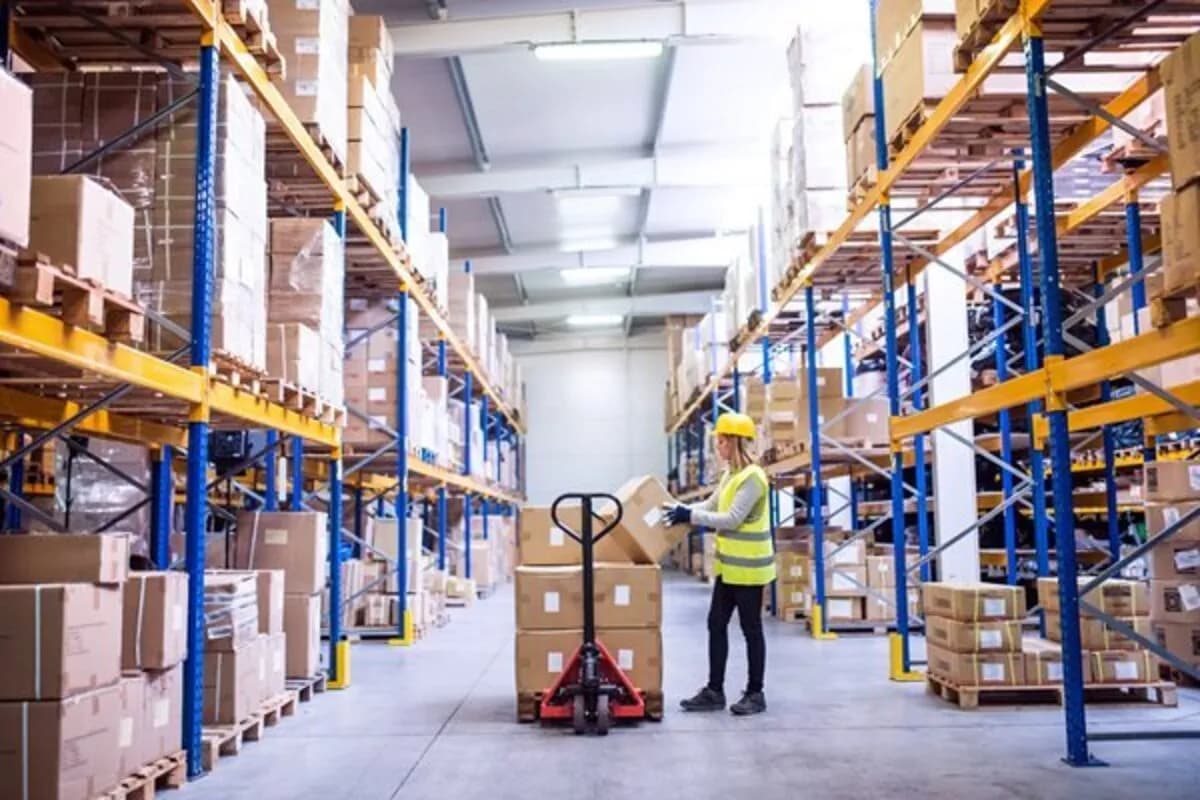 A warehouse aisle with racks of cartons and a worker moving a pallet