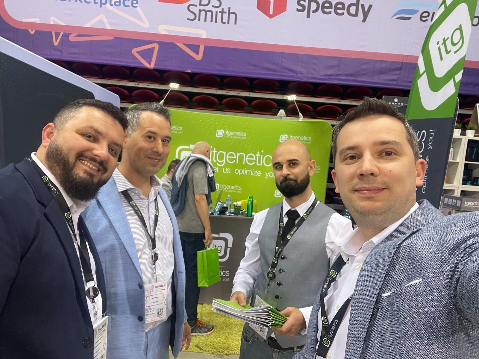 Ein Selfie von vier Personen mit itgenetics-Umhängebändern vor dem grünen itgenetics-Stand, eine von ihnen hält einen Stapel grüner Broschüren