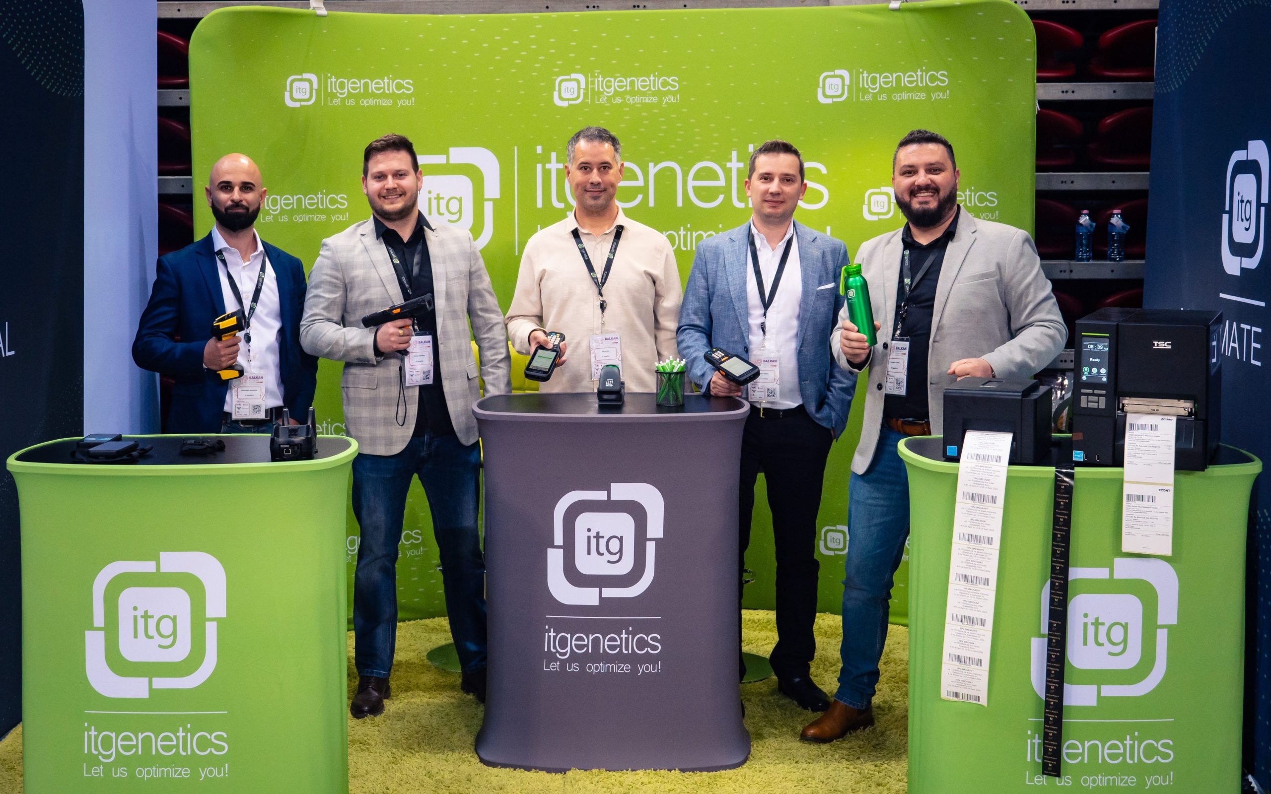 Fünf Personen posieren am itgenetics-Stand mit Handheld-Barcode-Scannern und mobilen Computern, neben Etikettendruckern und Streifen bedruckter Barcode-Etiketten